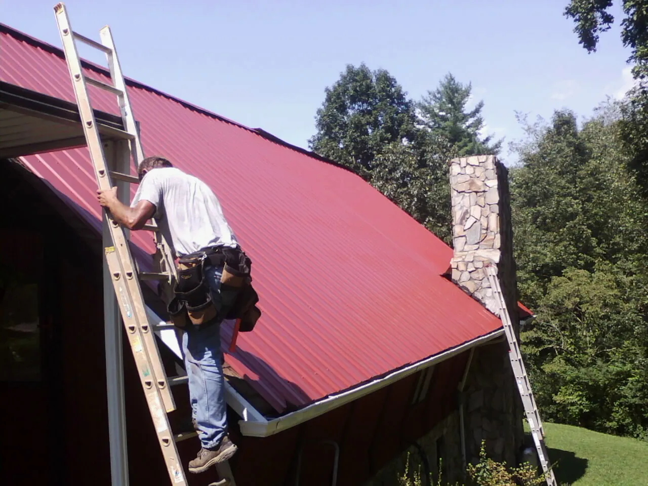 Roofer on a ladder beside a red metal roof and stone chimney — Soffit & Fascia Repair by Roofing Brothers in Jackson